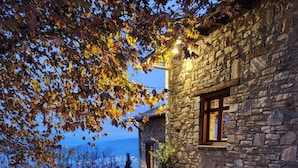 A stone building with a window and a door, a stone patio with scattered leaves, and a view of the sky and trees. - AURORA-INN (Volos)