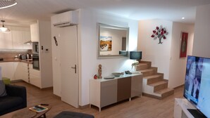 A modern living room with a sofa, ottoman, and a coffee table. There is a kitchen in the background with white cabinets and a dining area. - 4CH villa with pergola, swimming pool located 4km from Lac CARCANS and 9km from the Ocean (Carcans)