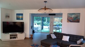 A living room with a flat-screen TV, a dark-colored sofa, and a large window. - 4CH villa with pergola, swimming pool located 4km from Lac CARCANS and 9km from the Ocean (Carcans)