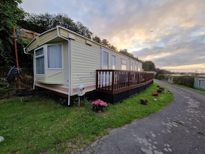 A mobile home with a deck, surrounded by greenery and a paved path. - 2 Bed Caravan With Aberystwyth Town Viev (Aberystwyth)
