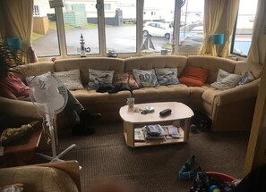Una sala de estar con un sofá seccional color beige, una pequeña mesa de centro y una lámpara de pie. Hay almohadas en el sofá y una ventana con vista a coches estacionados y un edificio.