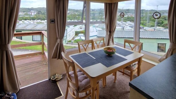 A dining area with a table and chairs, large windows with curtains, a view of a parking lot and buildings, and a ceiling light fixture. - 2 Bed Caravan - Sleeps 6 - Pets - Pool - Parking (Aberystwyth)