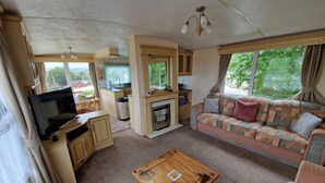 A compact living space with a TV, sofa, wooden coffee table, kitchenette, and a fireplace. - 2 Bed Caravan - Sleeps 6 - Pets - Pool - Parking (Aberystwyth)
