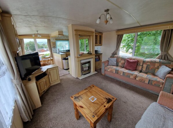 A compact living space with a TV, sofa, wooden coffee table, kitchenette, and a fireplace. - 2 Bed Caravan - Sleeps 6 - Pets - Pool - Parking (Aberystwyth)