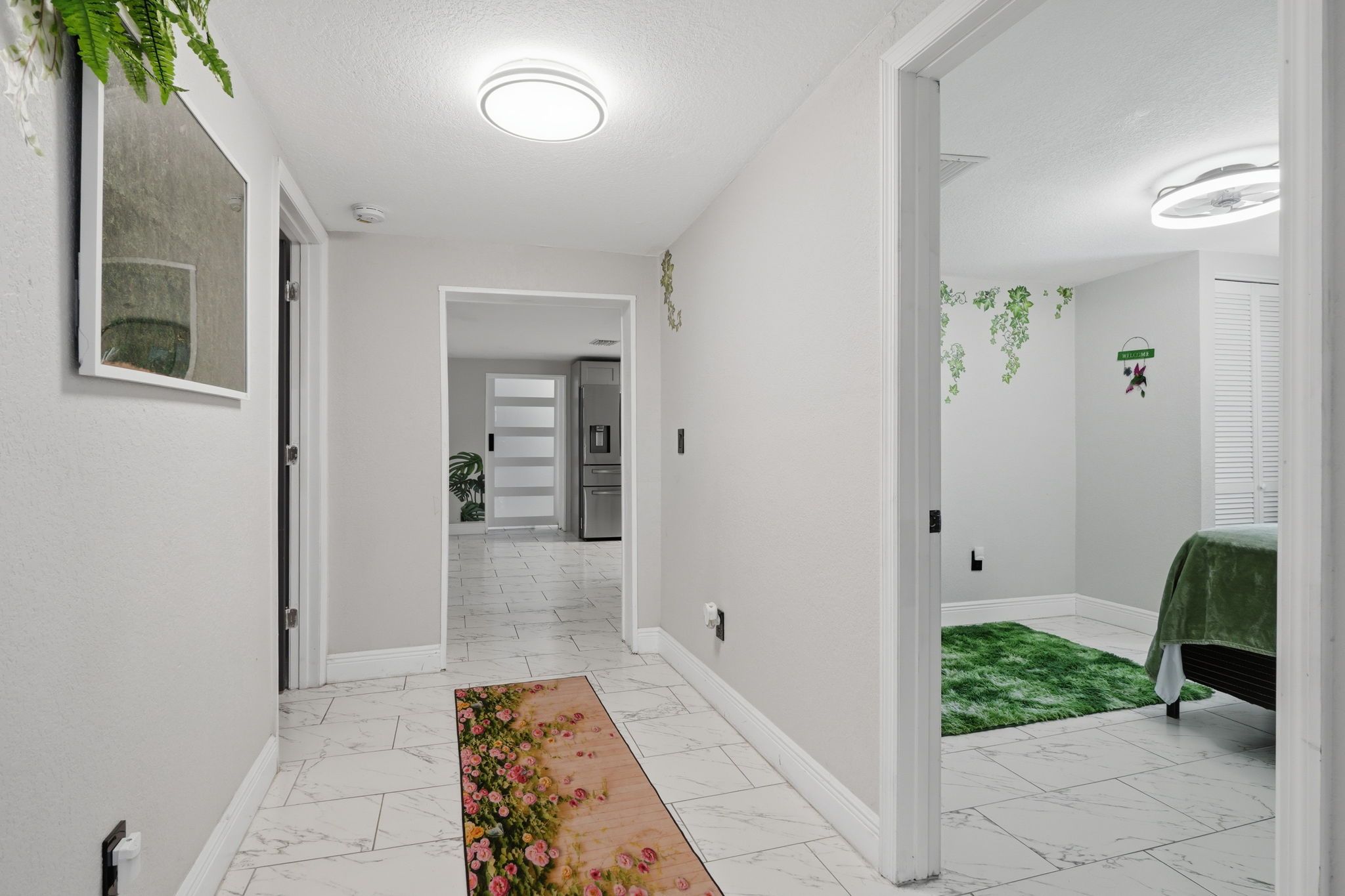 Un couloir doté d’un sol carrelé de carreaux blancs, d’un tapis à motifs floraux et d’une porte donnant sur une chambre où se trouve un tapis vert.