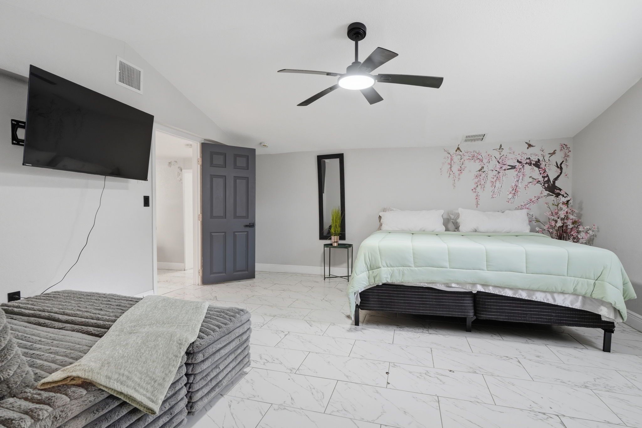 Une chambre à coucher comprenant un lit, un ventilateur de plafond, un téléviseur fixé au mur et une porte donnant sur une autre pièce.