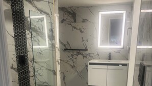 Une salle de bain dotée d’une douche à parois de verre, d’un sol en marbre blanc, d’un tapis de bain vert, d’un meuble-lavabo blanc avec un miroir et d’un mur en marbre orné d’un motif noir et blanc.