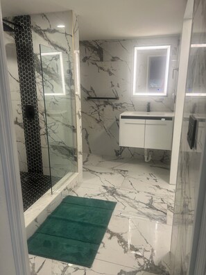 Un baño con cabina de ducha de vidrio, suelo de mármol blanco, alfombra de baño verde, tocador blanco con espejo y pared de mármol con diseño en blanco y negro.
