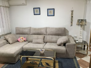 Een woonkamer met een beige bank, een glazen salontafel, een kamerkleed met een patroon en twee kunstwerken in lijsten aan de muur.