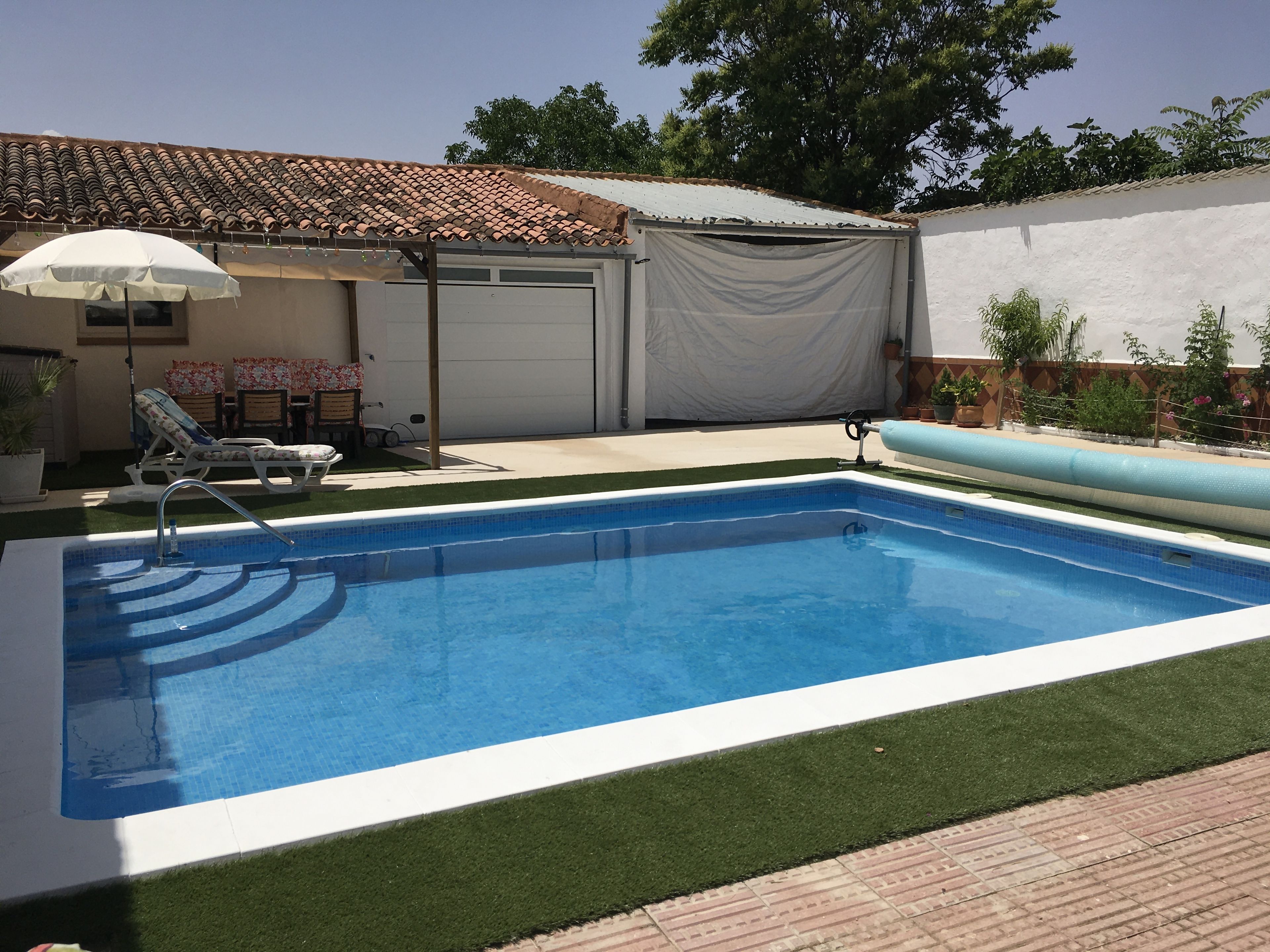 Une piscine avec un bord carrelé, un patio couvert comprenant une chaise et un parasol, et une porte de garage.