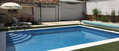 Una piscina con borde de azulejos, un área de terraza techada con una silla y una sombrilla, y una puerta de garaje.