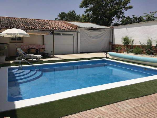 Une piscine avec un bord carrelé, un patio couvert comprenant une chaise et un parasol, et une porte de garage.