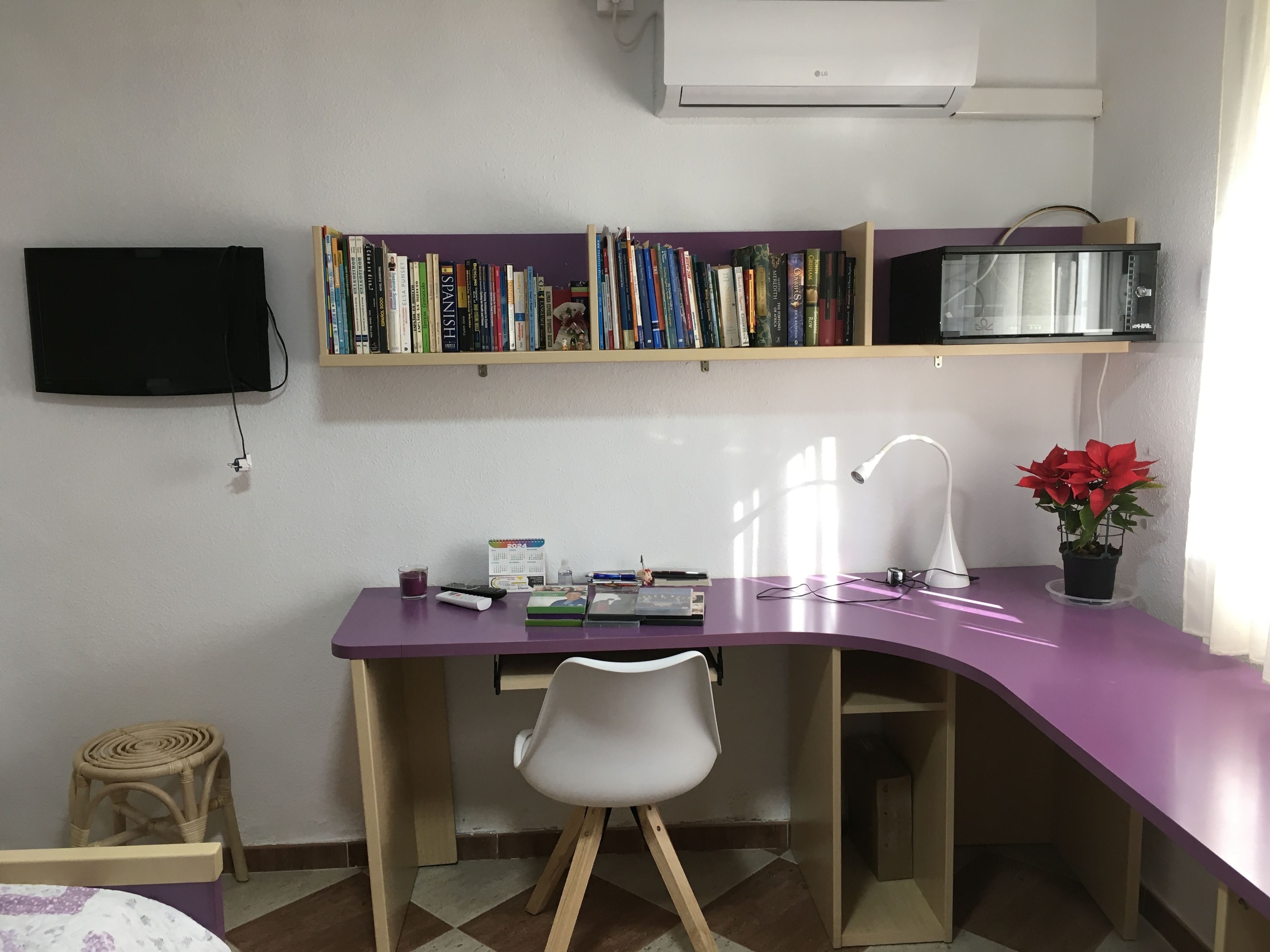 Un salón de estudio con un escritorio morado, una silla blanca, estanterías, un televisor, un microondas, una planta en maceta y un taburete pequeño.