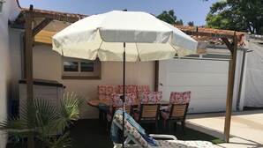 Een zonnig buitengebied met een witte parasol, een loungeset en een tafel met bloemenkussens.