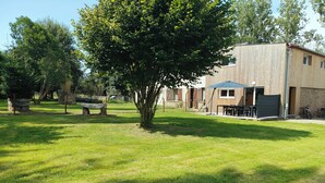Un espace extérieur verdoyant avec un arbre, un banc en pierre et un bâtiment doté d’une terrasse et de sièges extérieurs.