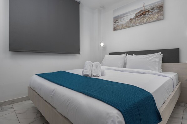 A neatly made bed with white linens and a teal throw, a headboard, and a painting of a lighthouse on the wall.