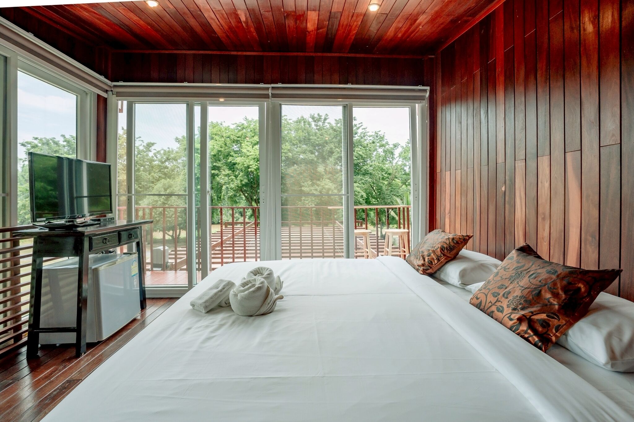 Une chambre spacieuse avec un grand lit, un bureau avec une télévision et un balcon offrant une vue sur les arbres.