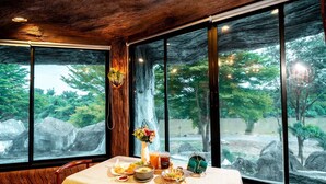 A dining table with a meal set, a vase with flowers, and a view of a natural landscape through large windows.