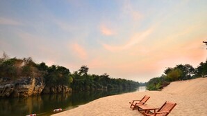 A sandy beach with three wooden lounge chairs and a calm river in the background.