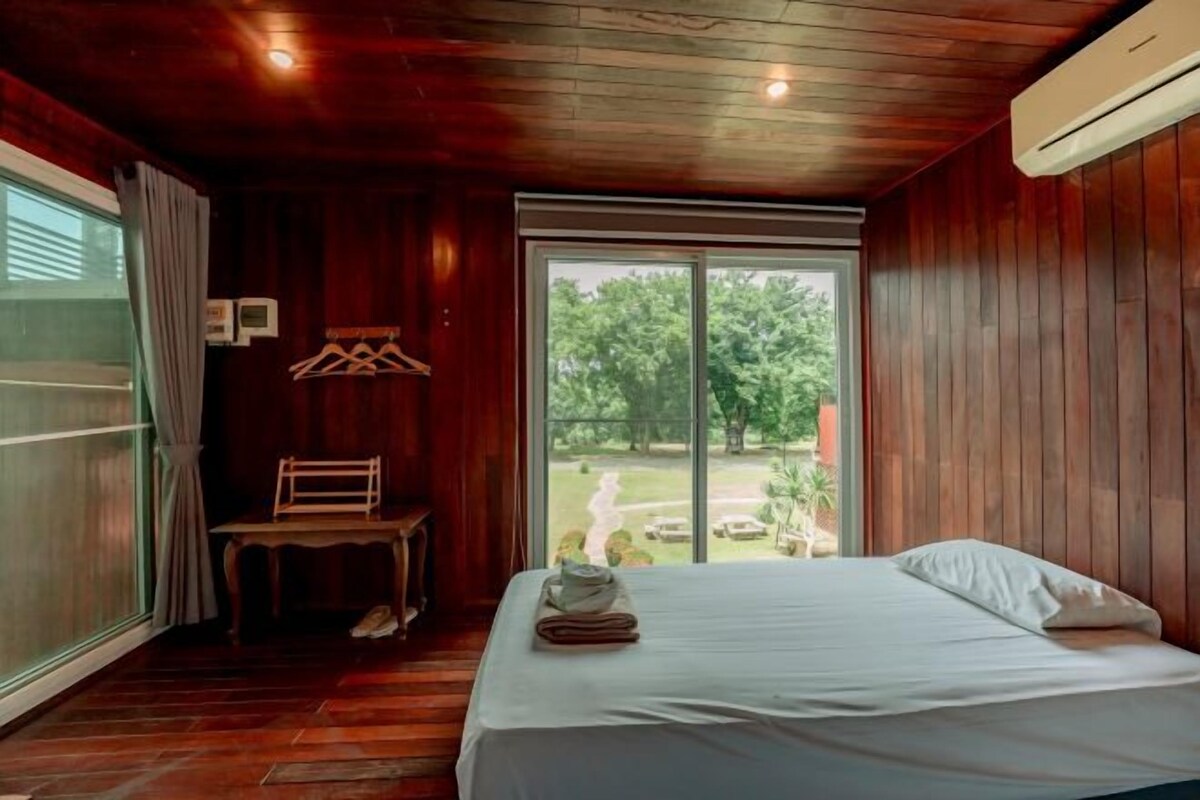 A bedroom with a wooden ceiling, a large window with a view of greenery, a bed with white linens, and a small table with a chair.