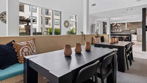 A modern dining area with a long table and chairs, a sofa with cushions, and a view of the building's exterior through large windows. - Bright 2BR Flat w/ Balcony + Pool | Walk to SoCo (Austin)