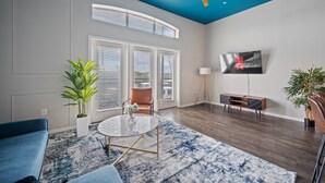 A modern living room with a blue ceiling, a TV mounted on the wall, a sofa, a coffee table, and two potted plants. - Modern 2BR Loft w/ Pool + Balcony in Heart of SoCo (Austin)