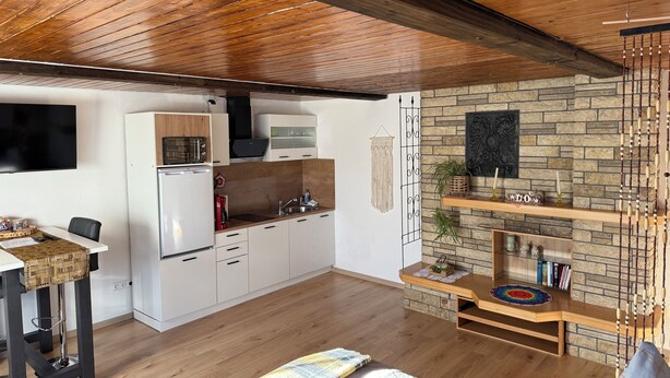 A modern kitchen with white cabinets, a microwave, and a refrigerator. There is a wooden ceiling and a stone wall in the background. - Cozy - Guest room (Notzingen)
