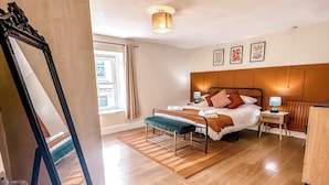 Une chambre d’hôtel avec un plancher en bois, un lit avec une tête de lit, une table de chevet avec une lampe, une fenêtre avec des rideaux et un miroir.