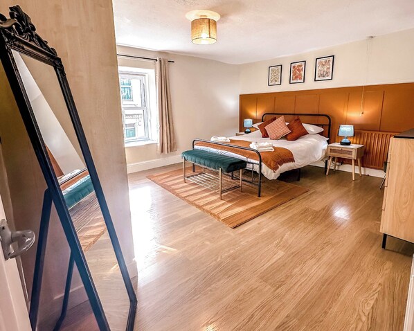 A hotel room with a wooden floor, a bed with a headboard, a bedside table with a lamp, a window with curtains, and a mirror.