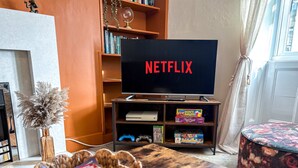Un salon avec un téléviseur affichant le logo « NETFLIX », une bibliothèque, une console de jeux et un pouf à motifs floraux.