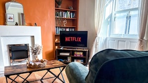 Un salon avec une cheminée, une bibliothèque, un téléviseur affichant le logo « NETFLIX » et une fenêtre avec des rideaux.