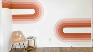 A room with a white wall featuring abstract orange and pink designs, a wooden floor, a beige chair, and a small white table. - The Retro duplex unit (Boise)