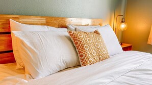 A neatly made bed with white linens and two pillows, one plain and one with a giraffe pattern, in a room with a wooden headboard and a nightstand with a lamp. - The Wildflower duplex unit (Boise)
