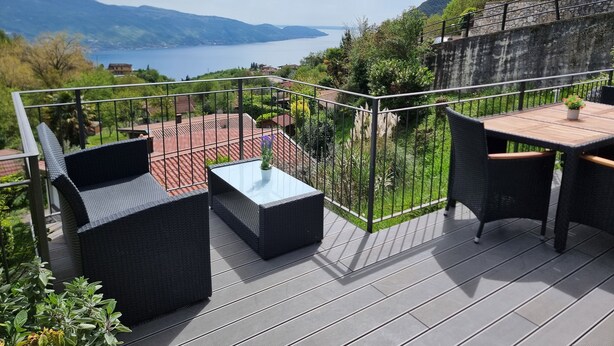Ein Balkon mit Korbmöbeln, einem kleinen Tisch mit Topfpflanze und Blick auf einen See und Berge.
