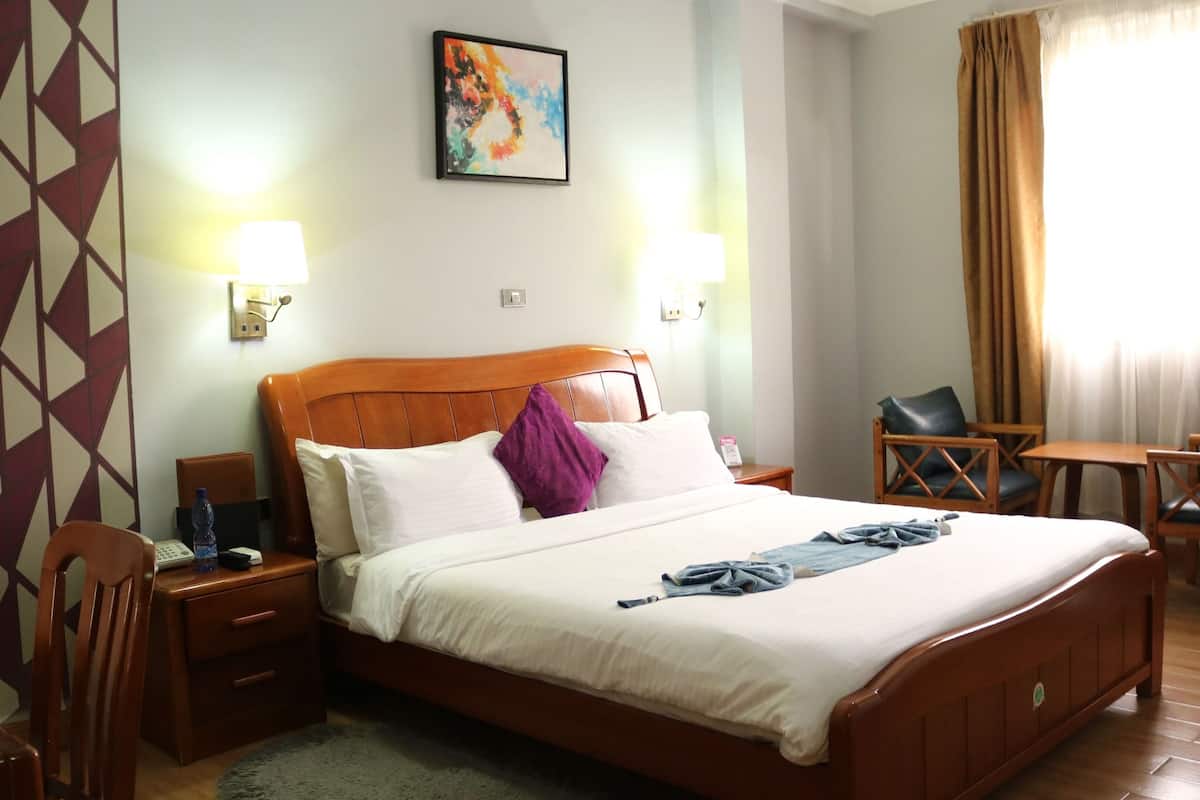 A hotel room with a wooden bed, two bedside tables, a chair, a small table, and a painting on the wall.