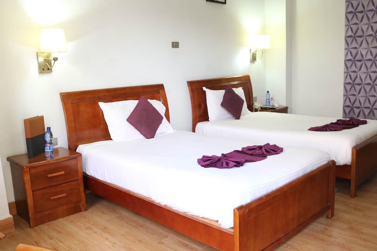 A hotel room with two wooden beds, white linens, and purple towels. There is a wooden nightstand with a water bottle and a lamp. The room has a wooden floor and a patterned wall.