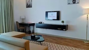 A modern hotel room with a sofa, a small table, a desk, a TV mounted on the wall, and a lamp.
