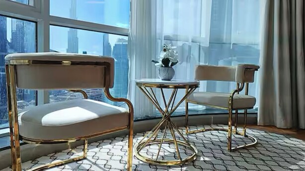 A modern room with a large window offering a city view, a round table with a vase of flowers, and two chairs with a gold frame.