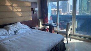 A hotel room with a large bed, a desk with a laptop, a chair, and a view of the cityscape through large windows.