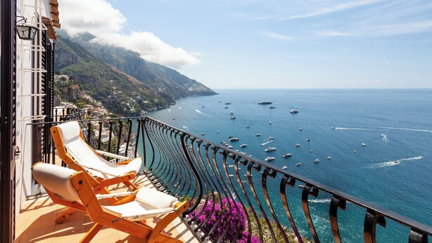 Un balcone con due poltrone che si affaccia su una vista costiera con barche e montagne.