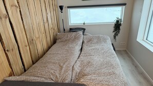 A single bed with a floral-patterned bedspread, a wooden headboard, a bedside lamp, a small potted plant, and a window with a view.
