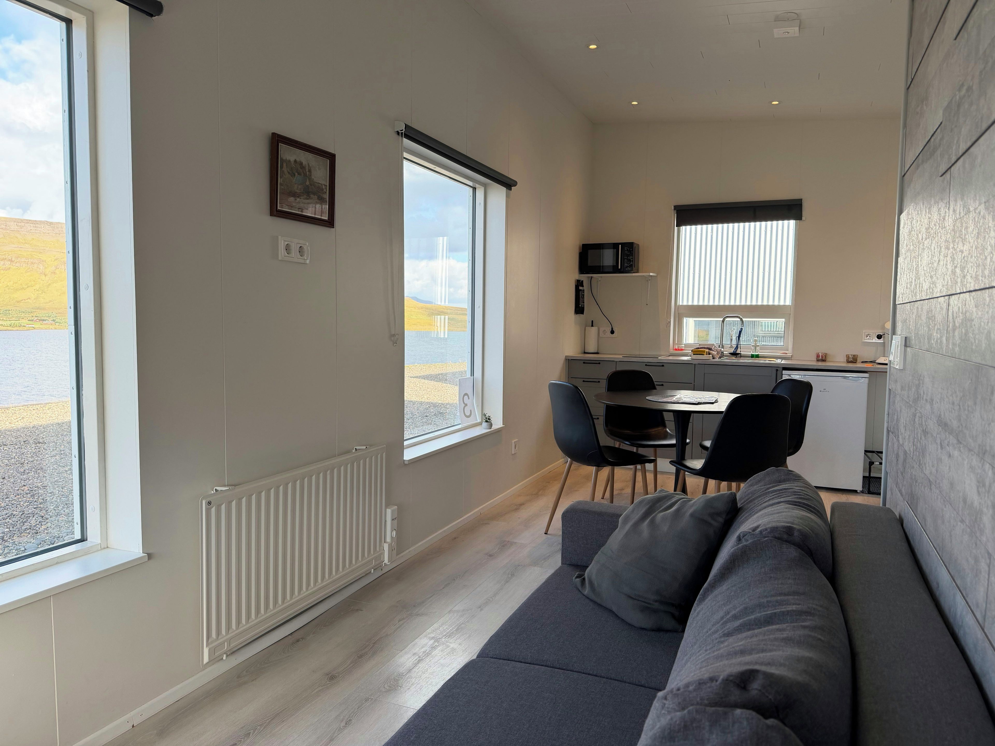 Un espacio habitable compacto con sofá, una mesa de comedor con sillas, una zona de cocina y grandes ventanas con vistas al exterior.