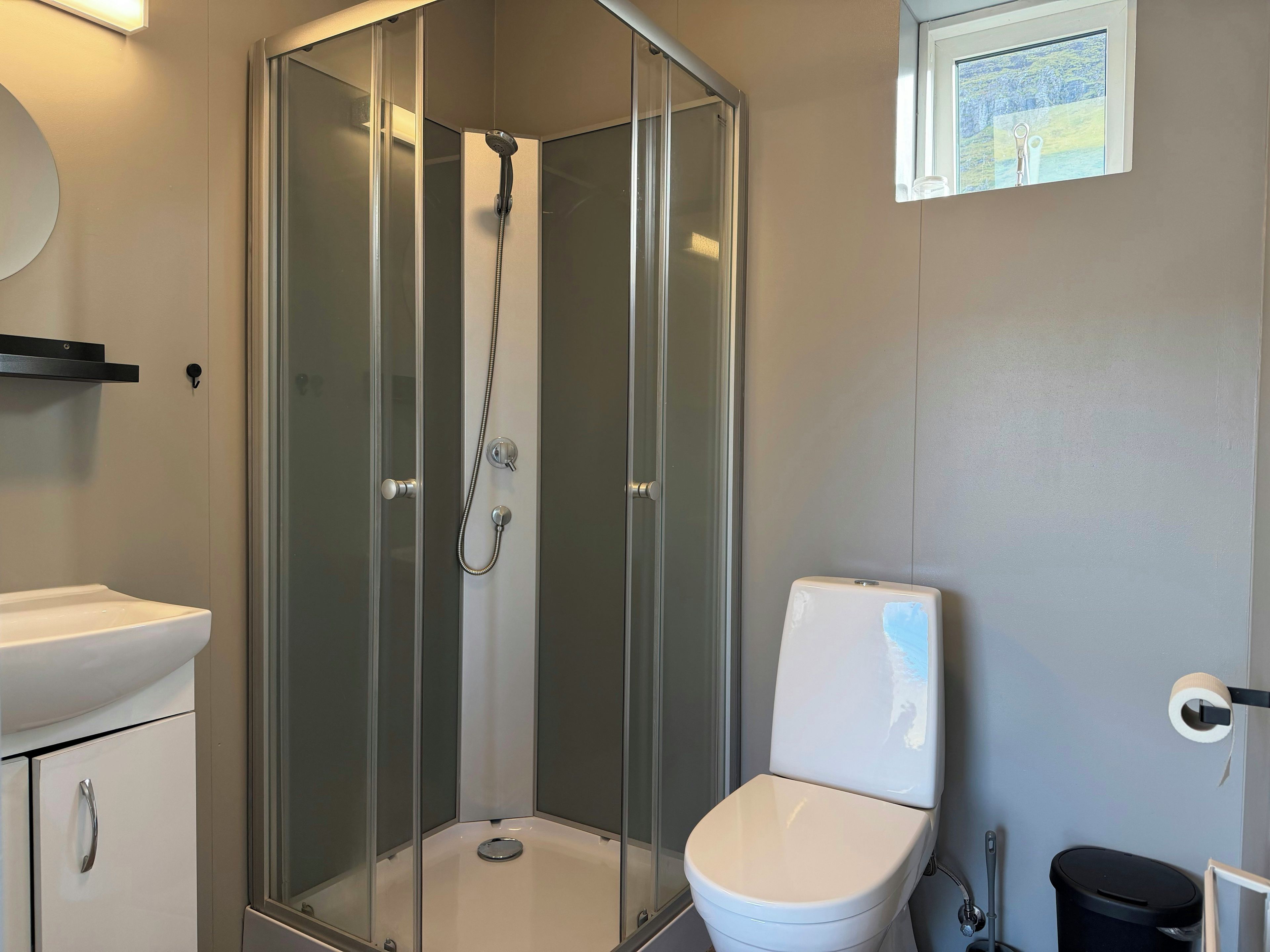 Un baño con ducha envuelta en vidrio, inodoro blanco, lavamanos y ventana con vistas.