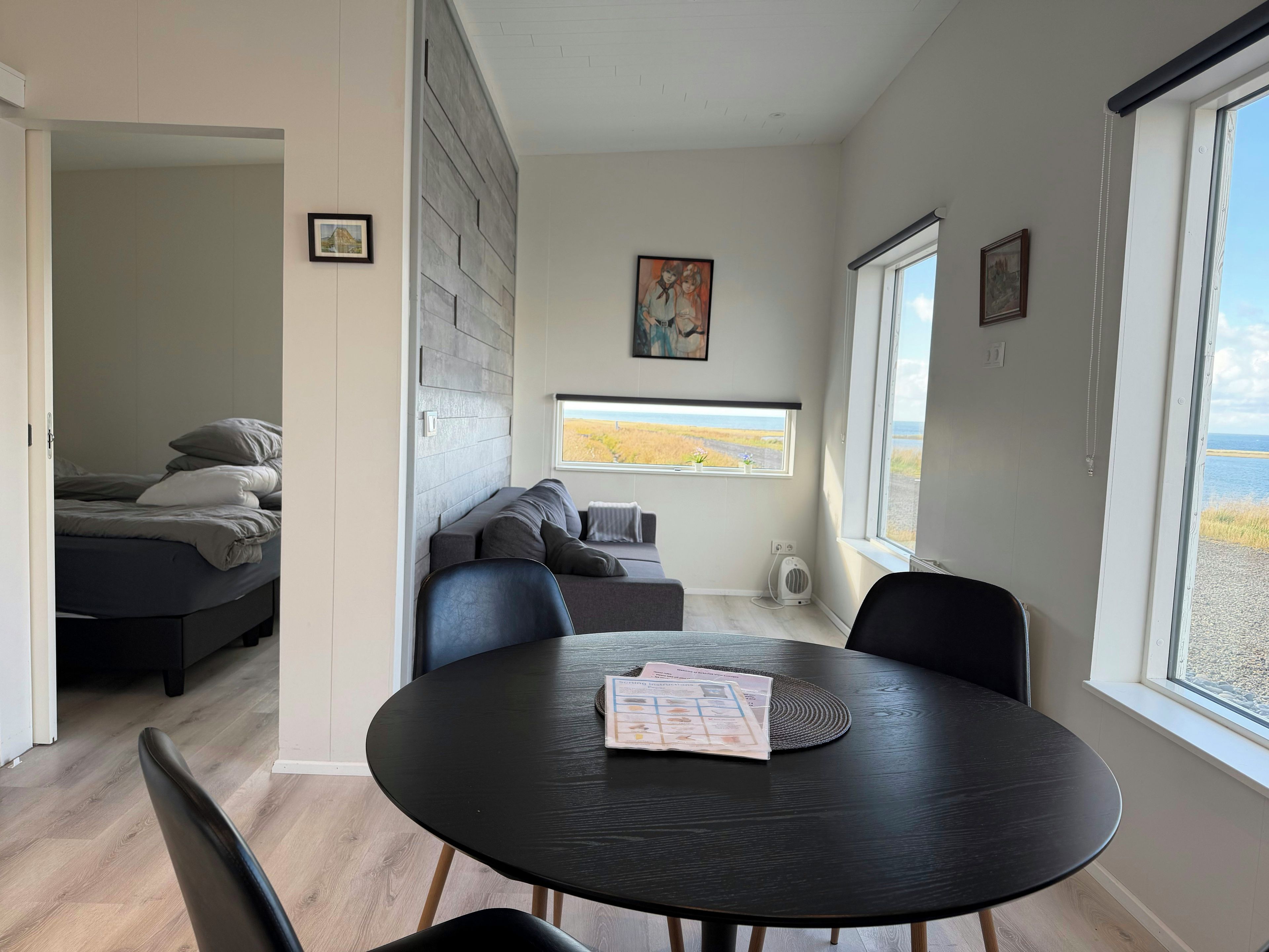 A modern room with a bed, a sofa, a dining table with chairs, and a large window offering a view of the beach.