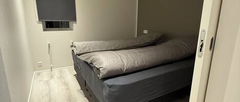 A small, single-bed room with a wooden floor, a wall-mounted TV, and a door to the right.