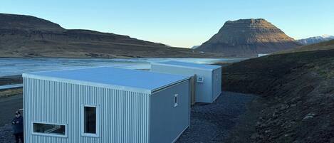 A row of corrugated metal buildings with a clear view of a mountainous landscape and a body of water in the background.