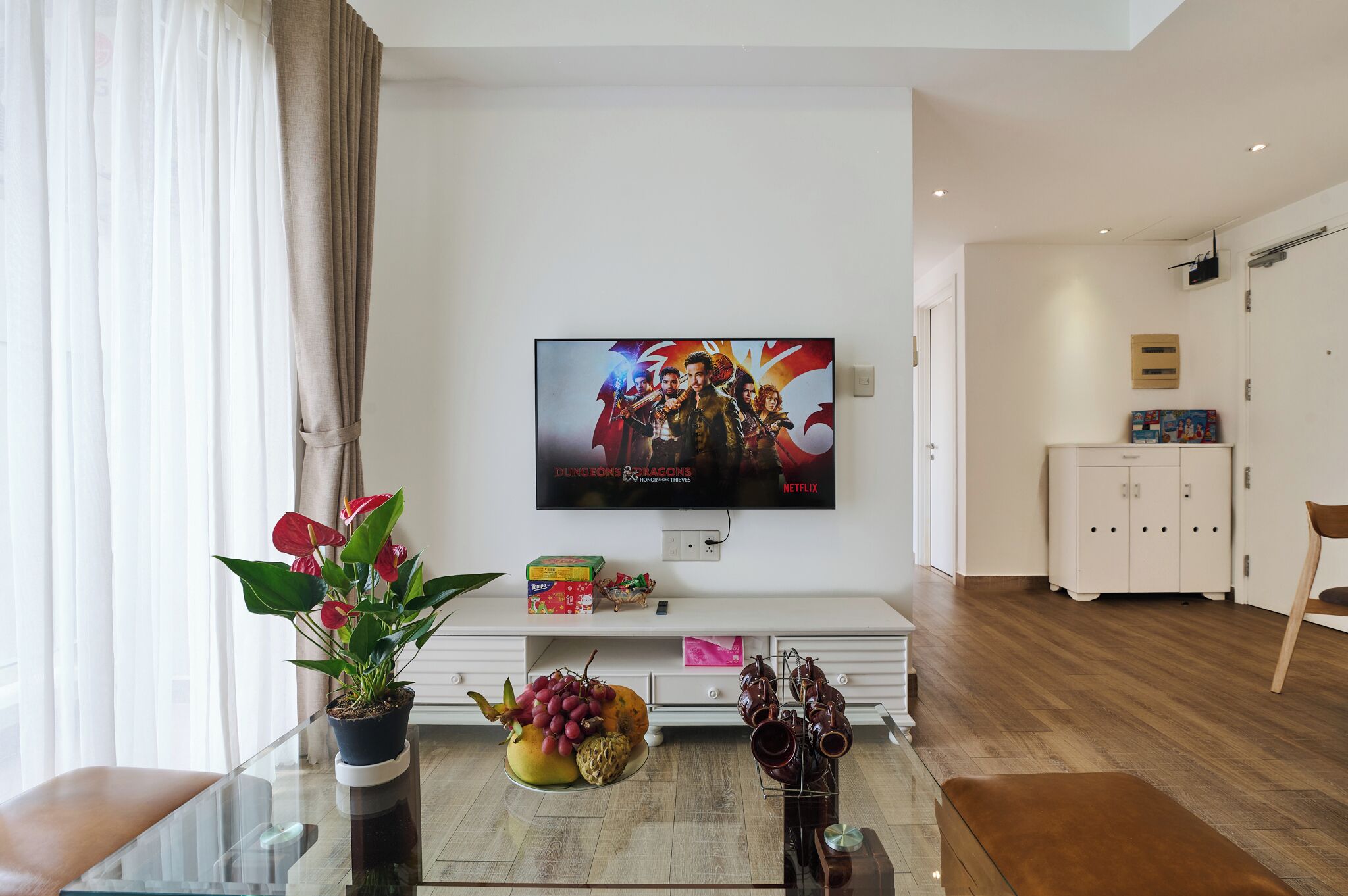A modern living room with a flat-screen TV displaying a movie, a glass coffee table with fruits and a potted plant, a white cabinet, and a wooden floor.