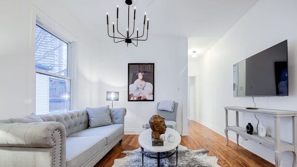A modern living room with a grey sofa, a round coffee table with a sculpture, a chandelier, a large TV, and a white console table. - Charming 2-bedroom, 1 bath apartment in historic Cabbagetown (Toronto)