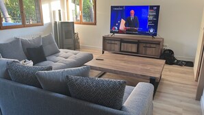 Ein modernes Wohnzimmer mit einem grauen Sofa, einem Holztisch und einem Fernseher, der eine Nachrichtensendung zeigt.