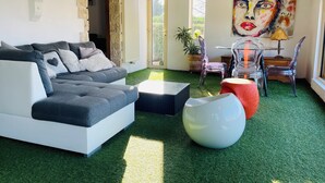 A modern living room with a sectional sofa, a glass coffee table, and a clear plastic chair. There is a large abstract painting on the wall and a potted plant near the window. - Crémieu, calm and relaxation, 2 steps from the center. (Crémieu)